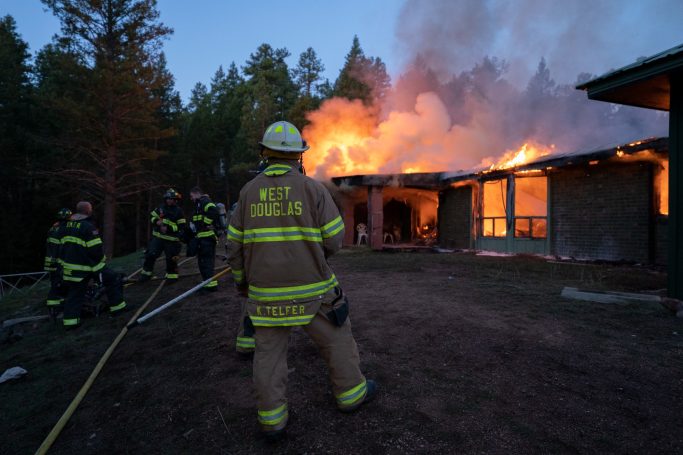 Firefighters battle a blaze at a building surrounded by smoke and flames in a forested area.