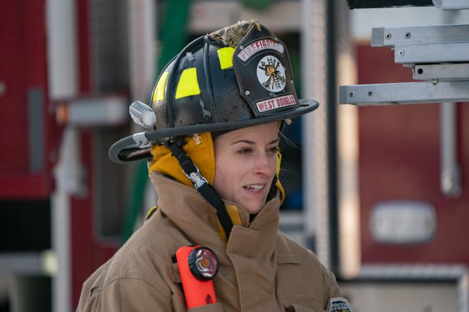 Female firefighter in gear, smiling, with a helmet and fire truck in the background.