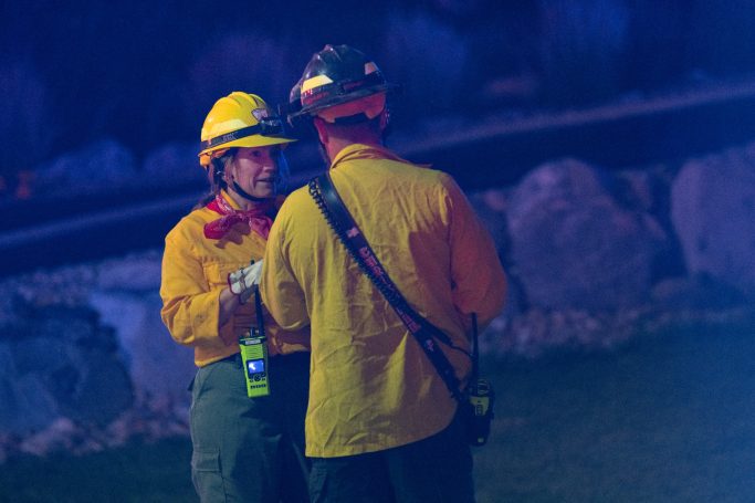 Two firefighters in yellow gear discuss a situation at night, illuminated by blue light.