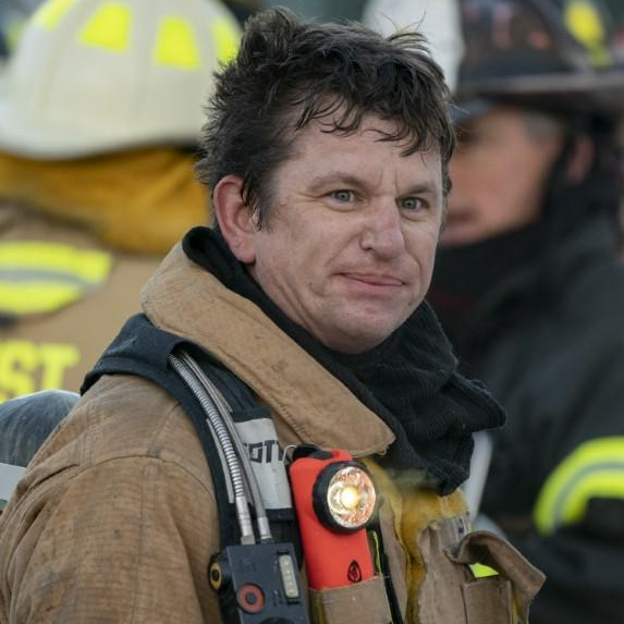 A firefighter in uniform with a flashlight, surrounded by colleagues in gear.