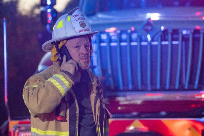 A firefighter in uniform talking on a phone with a fire truck in the background.