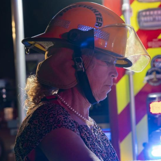 Woman wearing a firefighter helmet stands near emergency vehicles, focused and serious.