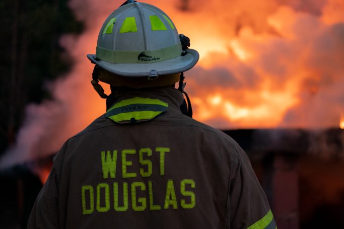 Firefighter in a protective helmet and jacket stands before a large blaze.