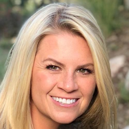 Smiling woman with long blonde hair outdoors, surrounded by greenery.