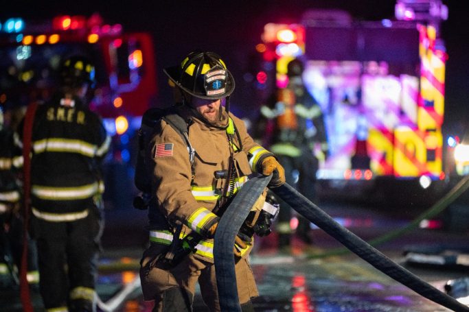 Firefighters respond to an emergency, handling hoses against a backdrop of emergency vehicles.