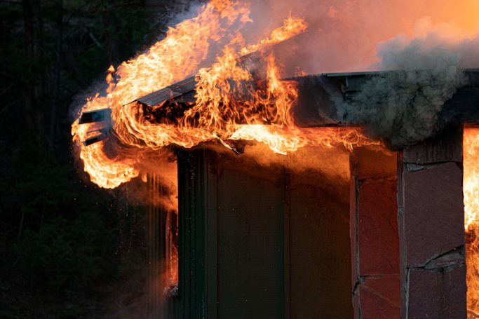 Flames engulf the edge of a building, with smoke rising against a dark background.