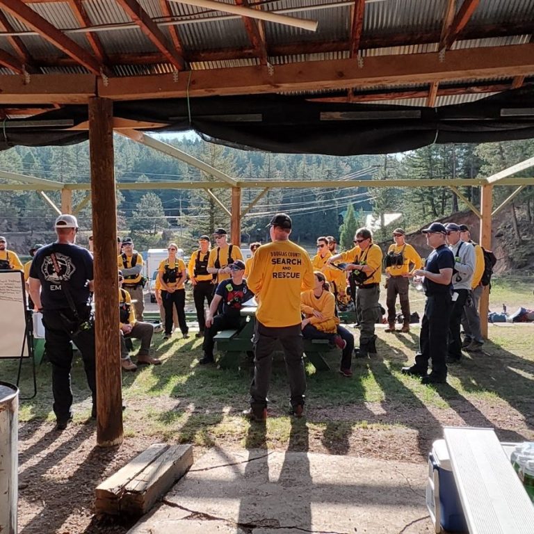 Group of people in yellow shirts gathered for a meeting outdoors in a wooded area.