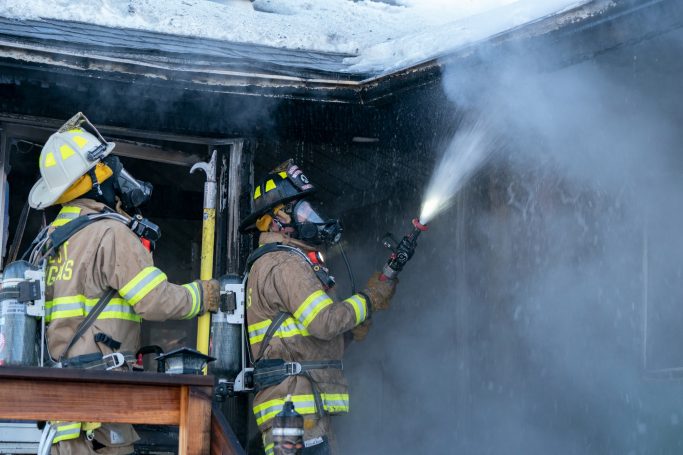 Two firefighters in protective gear extinguishing smoke from a building.