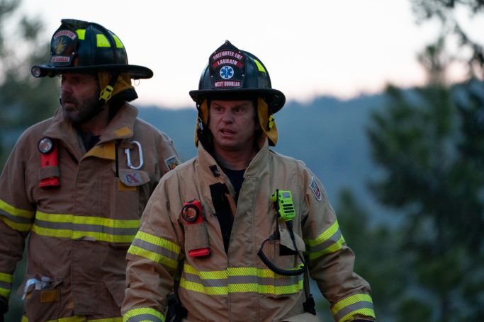Two firefighters in gear stand outdoors, surrounded by trees, with a soft sunset in the background.