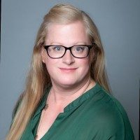 A woman with long blonde hair and glasses wearing a green blouse.