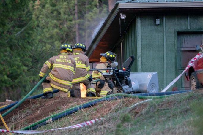 Firefighters in protective gear respond to a fire near a green building.