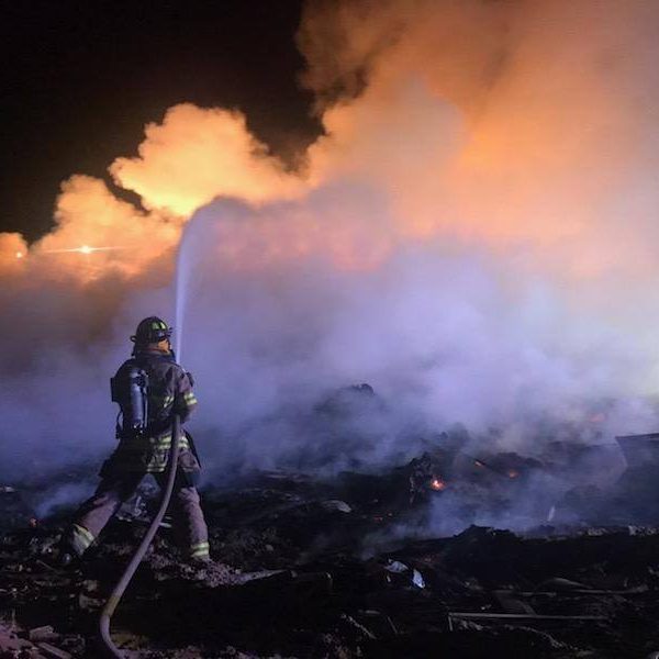 Firefighter spraying water on a blaze, surrounded by smoke and glowing flames at night.