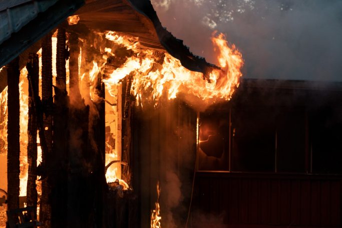 Flames engulfing the exterior of a wooden building, smoke billowing nearby.