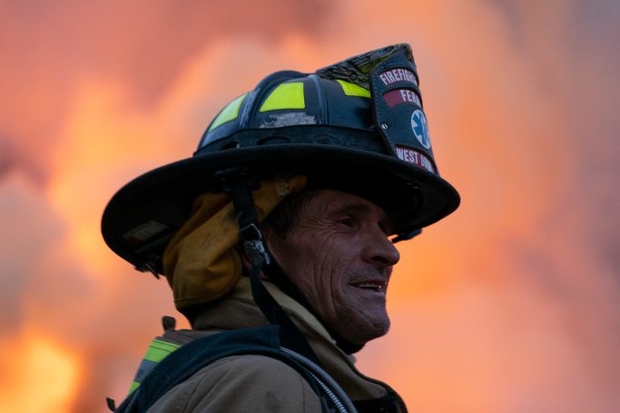 Profile of a firefighter with a helmet, smoke and flames in the background.
